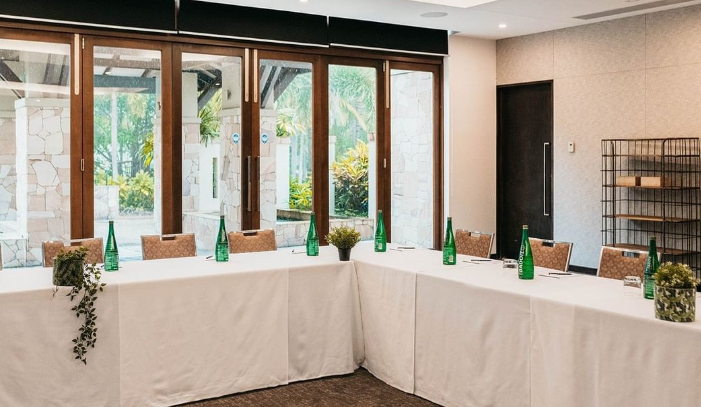 Looking into a conference room where the tables are arranged lengthwise and
into a corner shape. The tables are covered in a white drape and feature
several plants. A number of water bottles are also placed on the
table.
