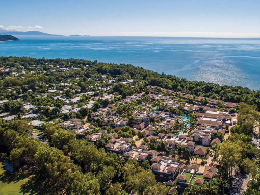 An aerial view of the seaside town of Port Douglas, showing suburban housing
complexes, with lots of greenery from the trees surrounding them. In the
distance is the blue ocean and some mountains on the left
horizon.